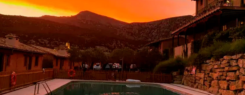alt="Piscina de alojamiento rural al atardecer con casas de piedra y montañas al fondo."